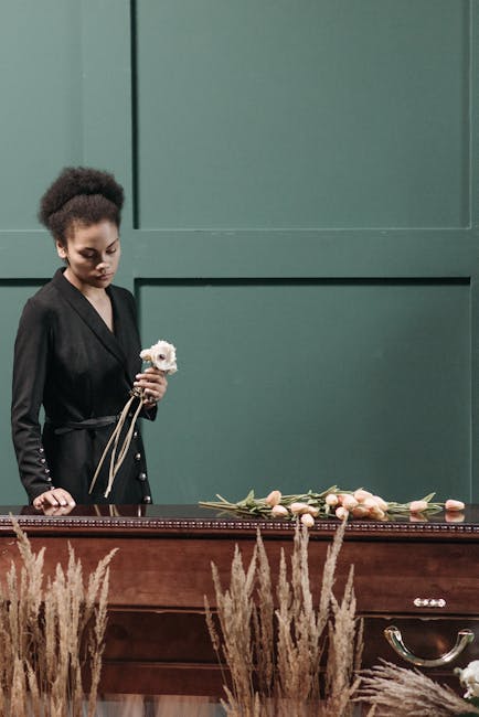 A young woman with dark, curly hair styled in an updo, dressed in a black blazer, stands beside a wooden coffin in a somber setting. She holds a single white flower in her right hand, with a gentle, c