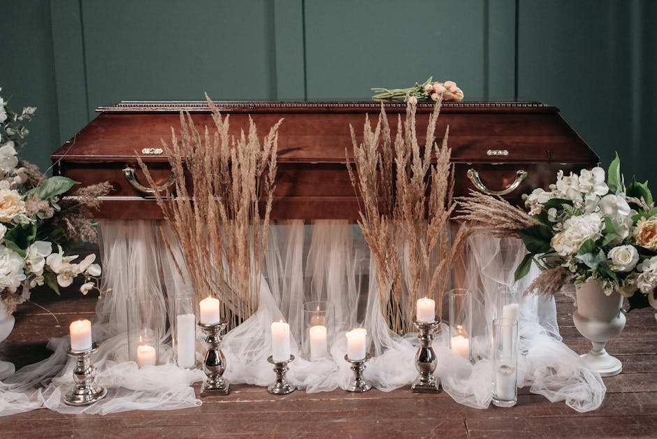 A somber floral arrangement centered around a polished wooden casket, draped with soft, white tulle fabric. Surrounding the casket are large white and cream-colored floral bouquets featuring roses, li